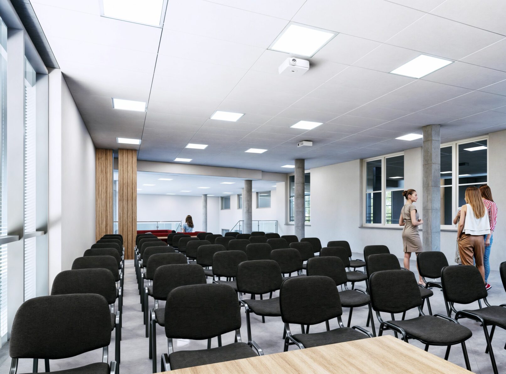 Rows of black upholstered chairs fill a large, bright multi-purpose room or lecture hall. Tall windows line the left wall, allowing natural light to enter. A projector is mounted on the white ceiling, which features square recessed lighting panels. Several people are standing or conversing in the background, near a wooden accent wall.